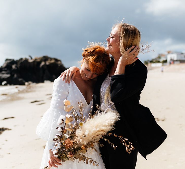 Two brides embrace on beach, one in white dress holding dried flower bouquet and other bride in black suit with silver sequin top