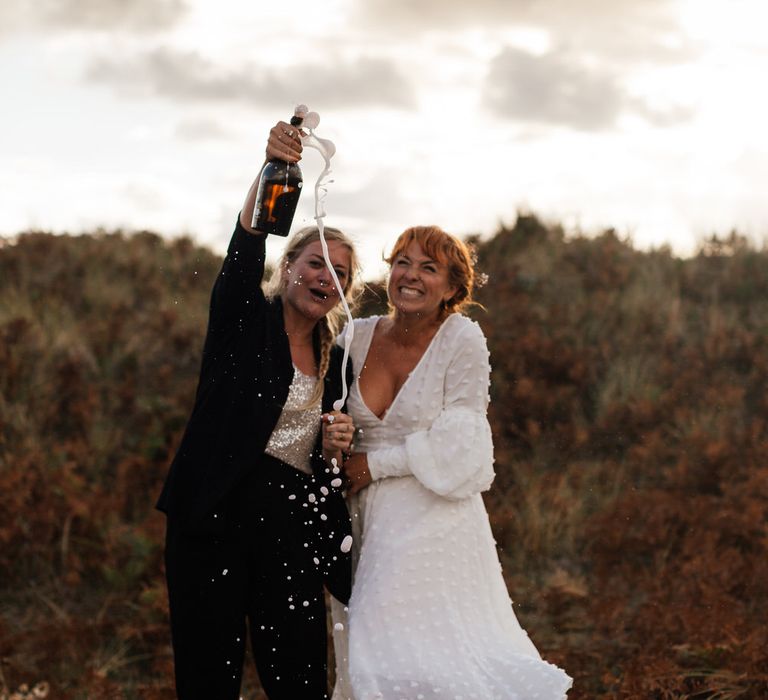 Bride in black suit spills champagne as bride in white dress laughs on beach