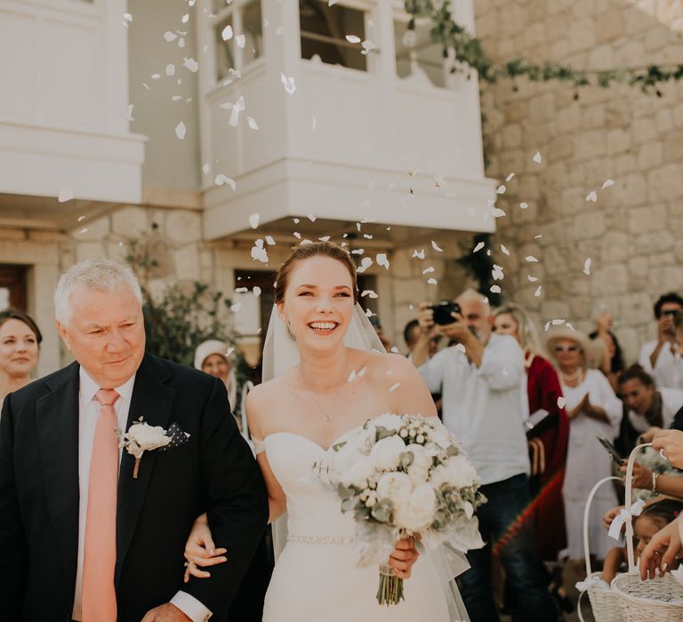 Bride and her father walking up an aisle of guests throwing white confetti 