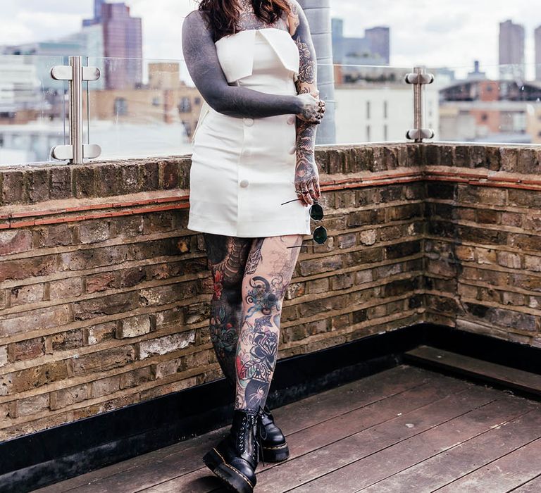 Tattooed bride in a short wedding dress, Dr Marten boots and appliqué headband standing on a roof terrace 