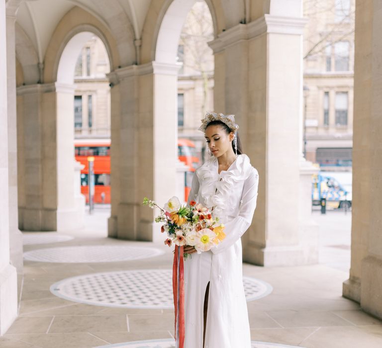 Bride wears golden crown with ruffled front wedding gown whilst holding bright and bold floral bouquet tied with pastel velvet ribbon