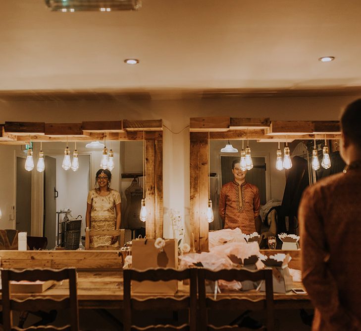 An Asian bride wears a gold Lehenga as she stands next to her new husband looking into the mirror.