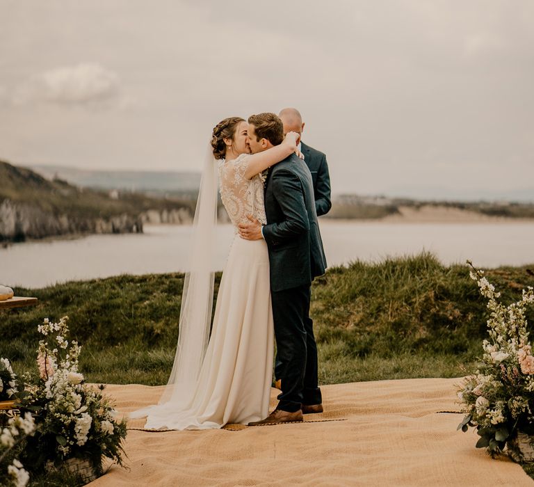 Smiling bride in lace top with capped sleeves and satin skirt has first kiss with groom in navy suit at clifftop ceremony for Dunluce Castle wedding