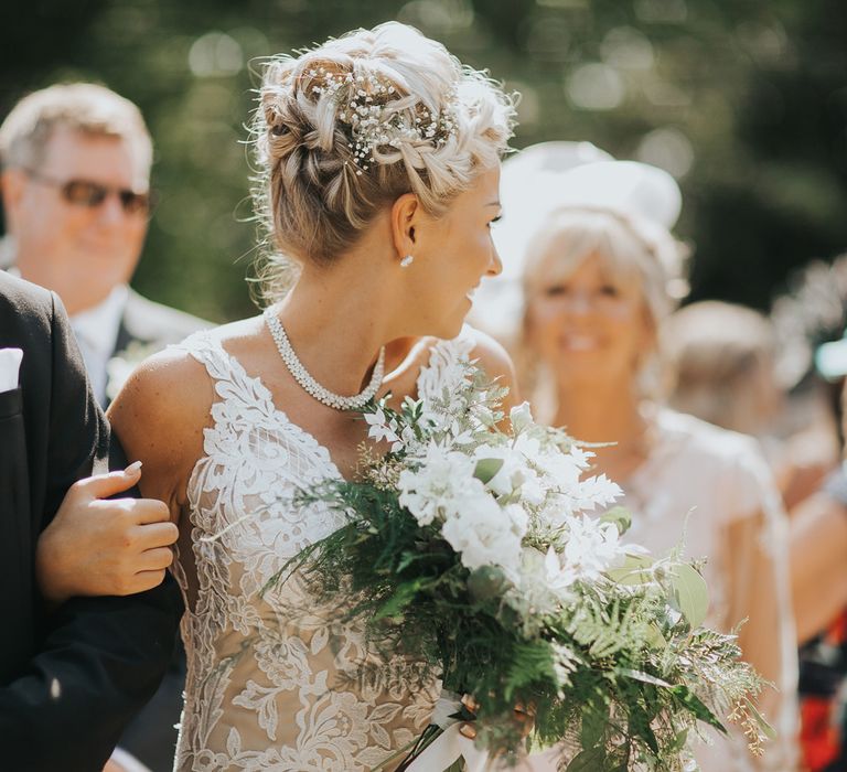 A bride in a lace wedding dress carries a bouquet as she walks down an outdoor aisle. Her blonde hair is up high with a braid and gypsophelia in the side.