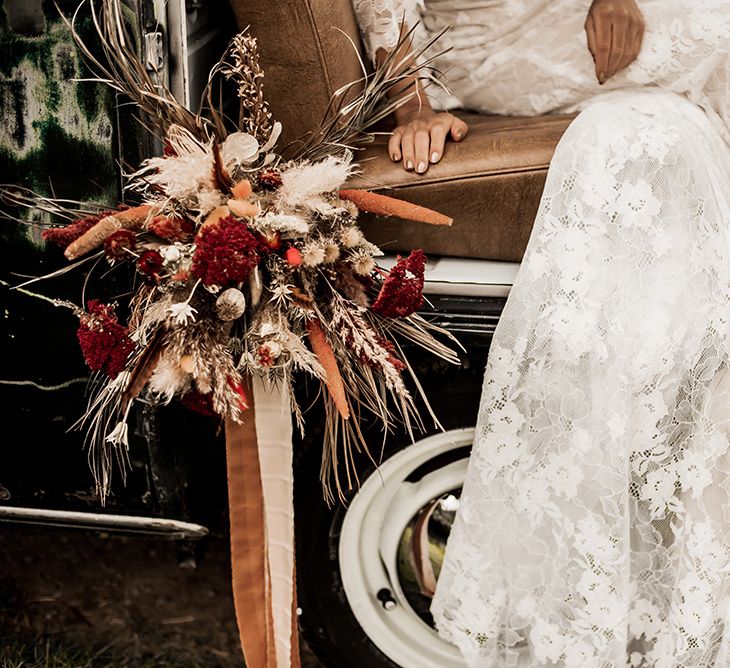 Bride in a lace wedding dress sitting in a VW Camper van with her red and dried flower bouquet 