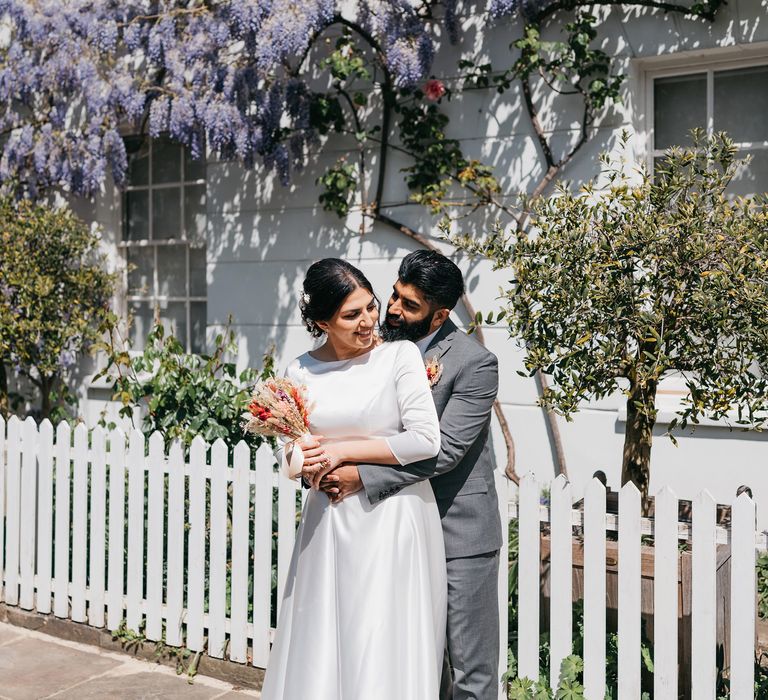 Groom wraps his arms around his bride in the sunshine as they stand in front of white picket fence and house with hanging purple wisteria tree in the garden