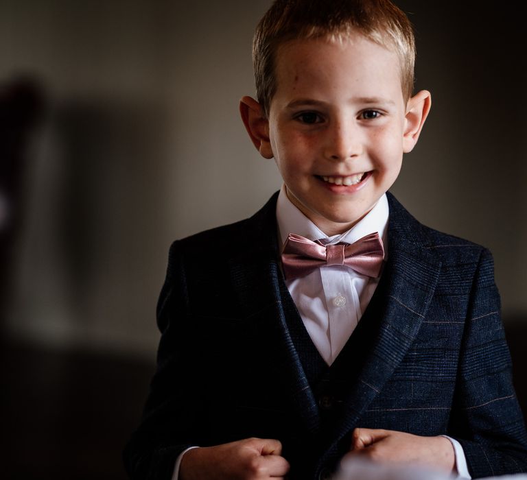 Young groomsman in dark blue checked suit, white shirt and pink silk bowtie smiles at the camera before wedding at The West Mill Derby