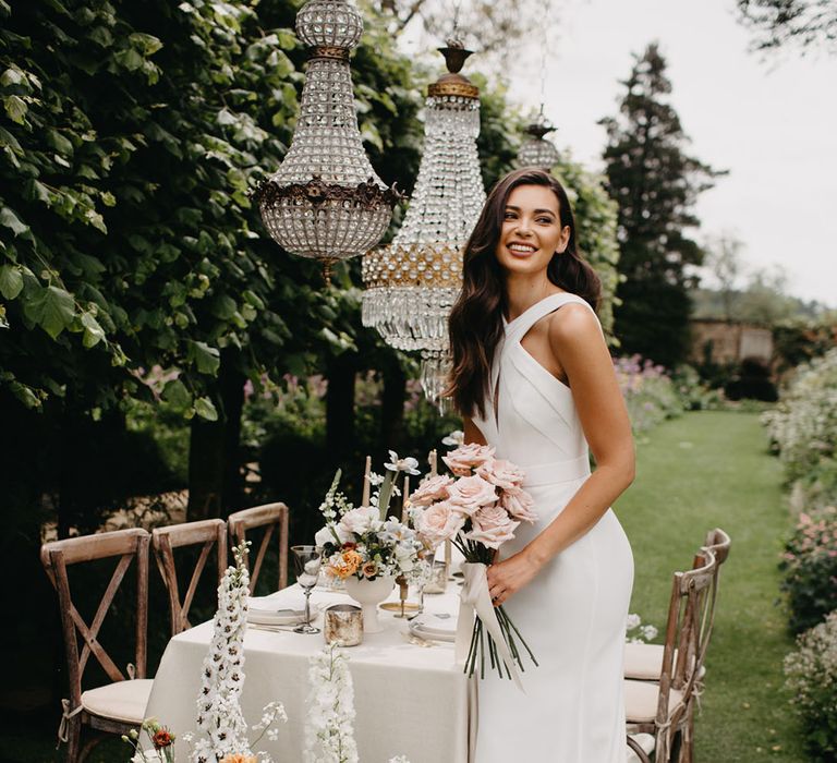 Bride in an A Line halter neck Suzanne Neville wedding dress standing in front of an elegant outdoor reception with hanging chandeliers 