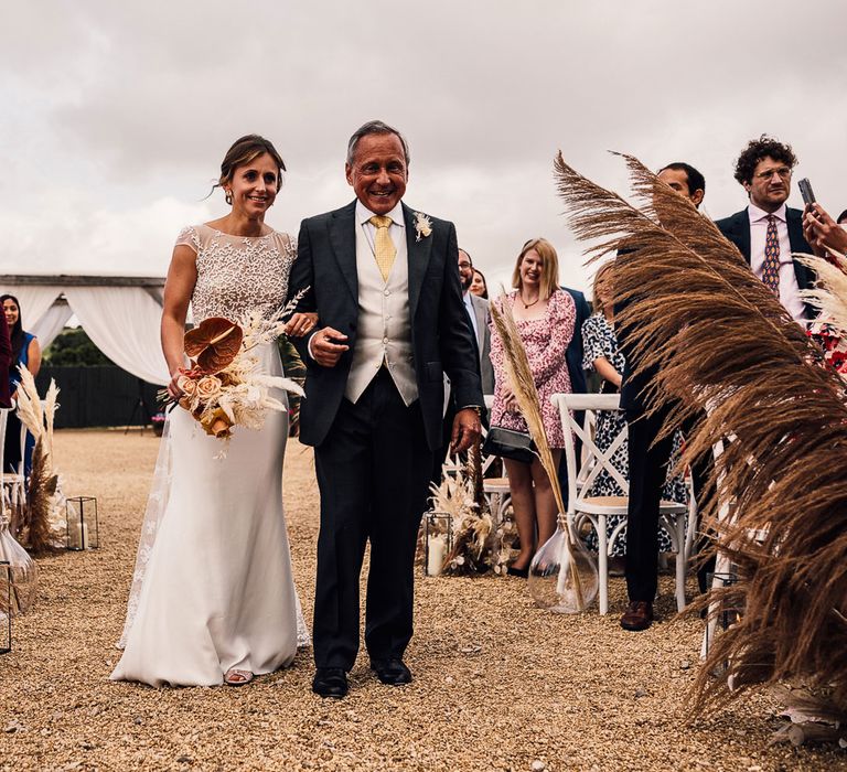 Bride in white Rime Arodaky wedding dress holding rustic dried bridal bouquet walks down the aisle arm in arm with man in blue suit and yellow tie during outdoor wedding ceremony in Dorset with boho decor