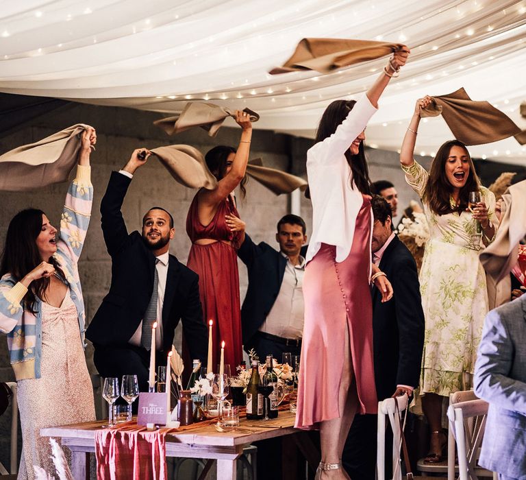 Wedding guests stand on chairs and wave napkins at boho wedding breakfast for summer wedding in Dorset