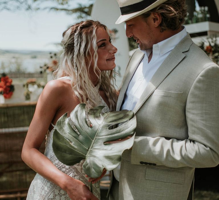 Bride in white halter neck lace boho wedding dress and tropical flower crown smiles at groom in linen suit and white straw fedora whilst holding palm leaf as they stand in front of campervan cocktail bar for birds of paradise wedding inspiration