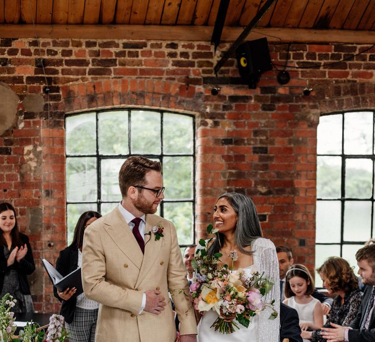 Groom in double breasted linen suit and gold Nike Air Force 1 trainers holds hands with bride in satin Halfpenny London wedding dress and tasselled bridal cape holding multicoloured bridal bouquet during wedding ceremony at Loft Studios London