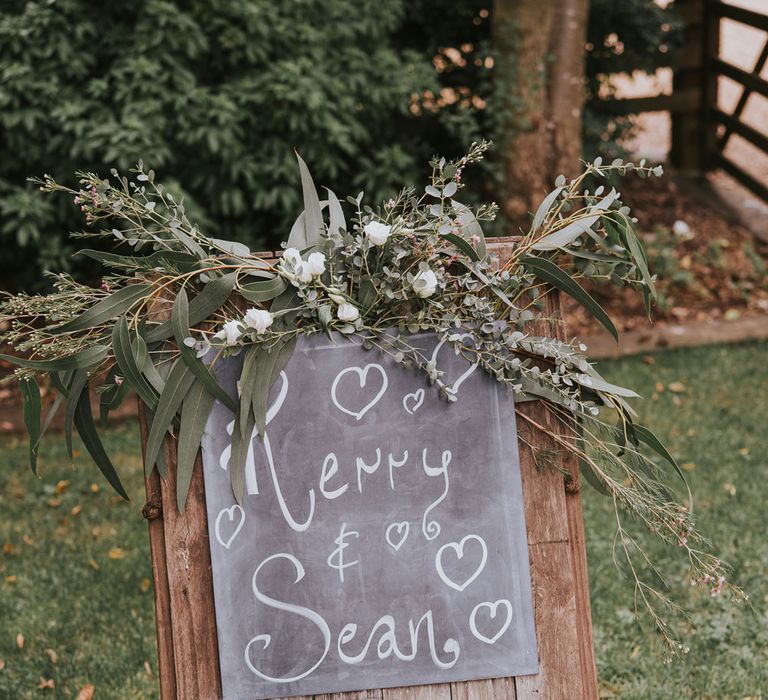 Wood and chalkboard wedding sign reading 'Kerry & Sean' topped with white florals and green foliage at Tythe Barn wedding with barn wedding flowers