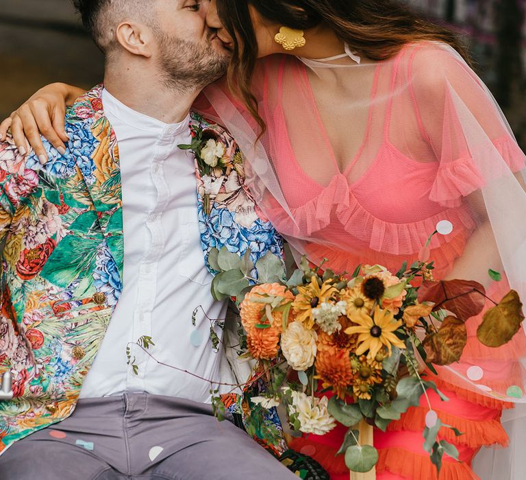 Bride n a coral wedding dress and cape veil and flower crown kissing her groom in a white shirt and brightly coloured patterned jacket 