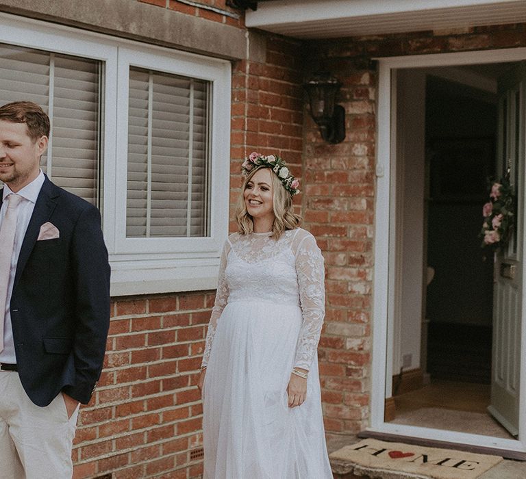 First look with pregnant bride in an empire line wedding dress with lace ling sleeves and pink flower crown approaching her groom in shorts and a blazer 