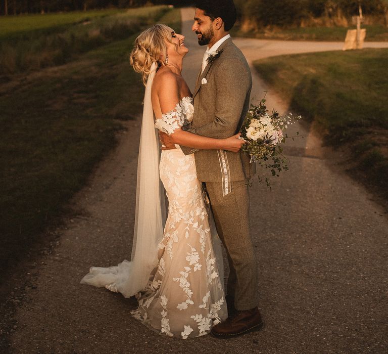 Bride in white Enzoani wedding dress and veil holding white and pink bridal bouquet hugs groom in grey suit as they stand on country lane during golden hour at Inkersall Grange Farm wedding