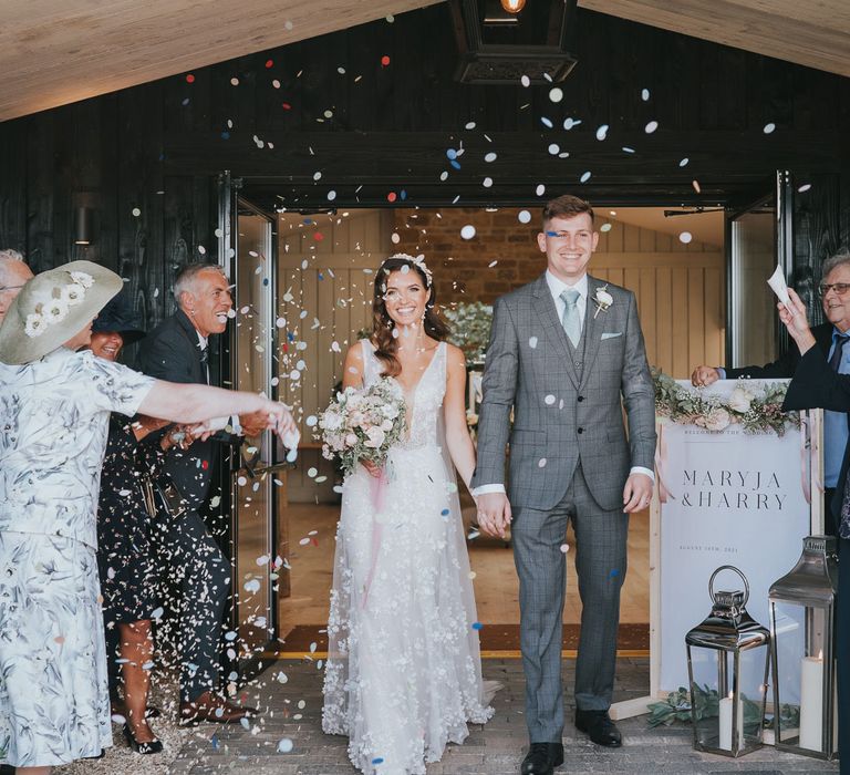 Bride in applique Berta wedding dress and headband holding white and pink bridal bouquet holds hands with groom in grey checked three piece suit as they walk out of barn whilst guests throw confetti for summer wedding at Primrose Hill Farm