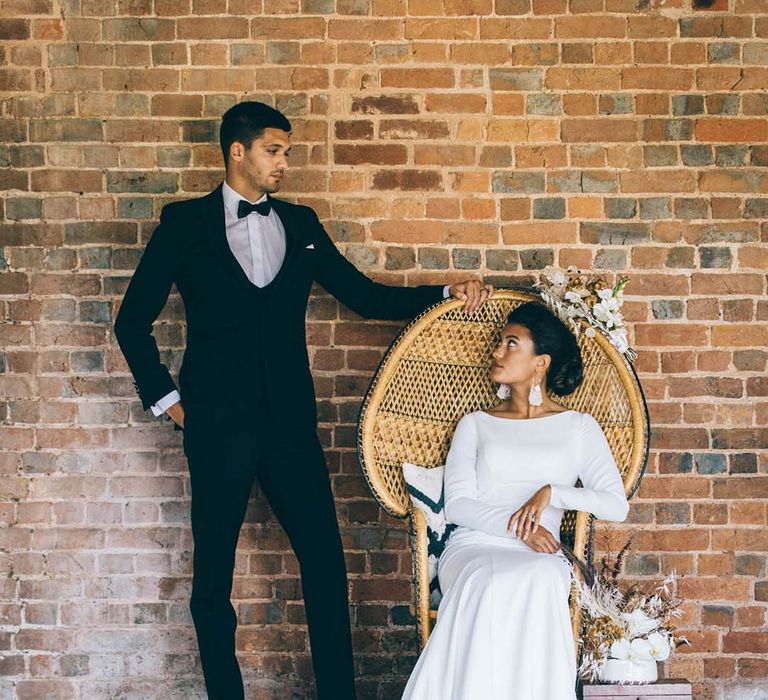 boho black tie wedding at Brickhouse vineyard with groom in a tuxedo and bride in a minimalist wedding dress sitting in a wicker peacock chair 