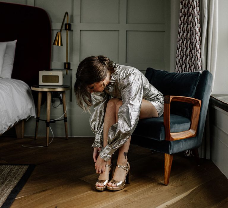 Bride wearing platform gold heels, long detailed sleeved short gold wedding dress in a room with panelled walls & a retro radio 