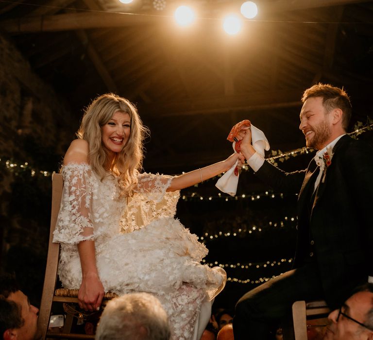 Laughing bride holds hands with smiling groom during horah at evening wedding ceremony under fairy light canopy