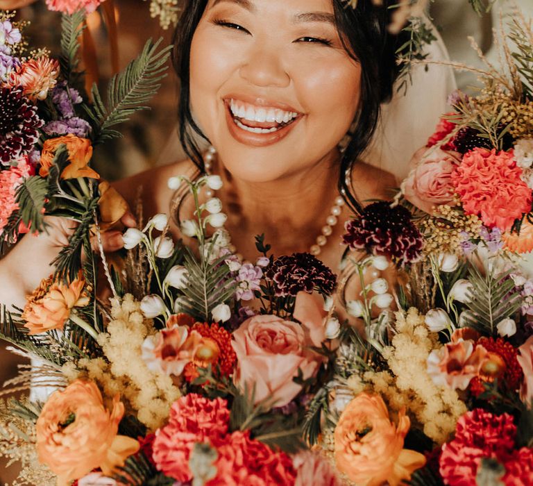 Bride with curled updo and pearl necklace smiles through hoop of colourful mixed florals for wedding at Scale in Liverpool