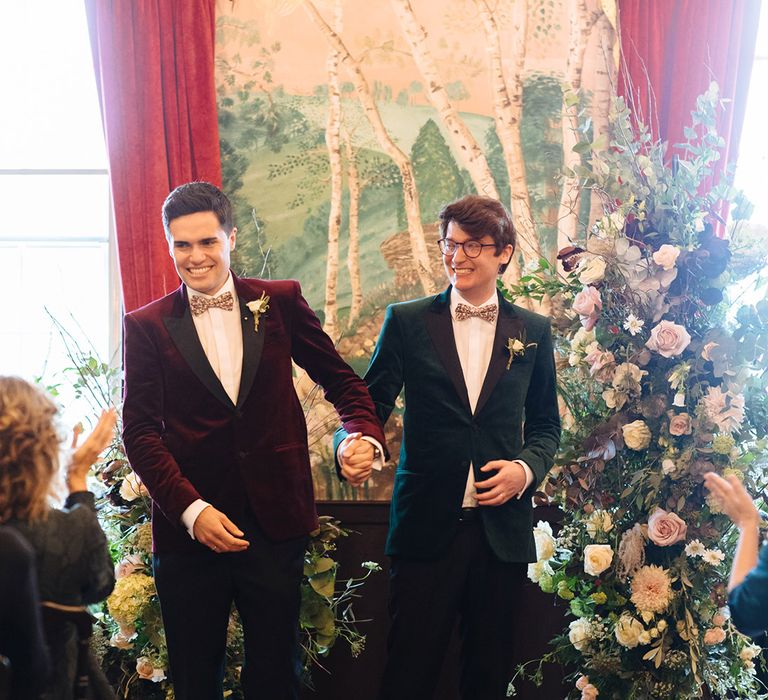 Grooms in burgundy and forest green velvet tuxedo jackets with matching floral bow ties at quirky wedding venue with forest wallpaper 