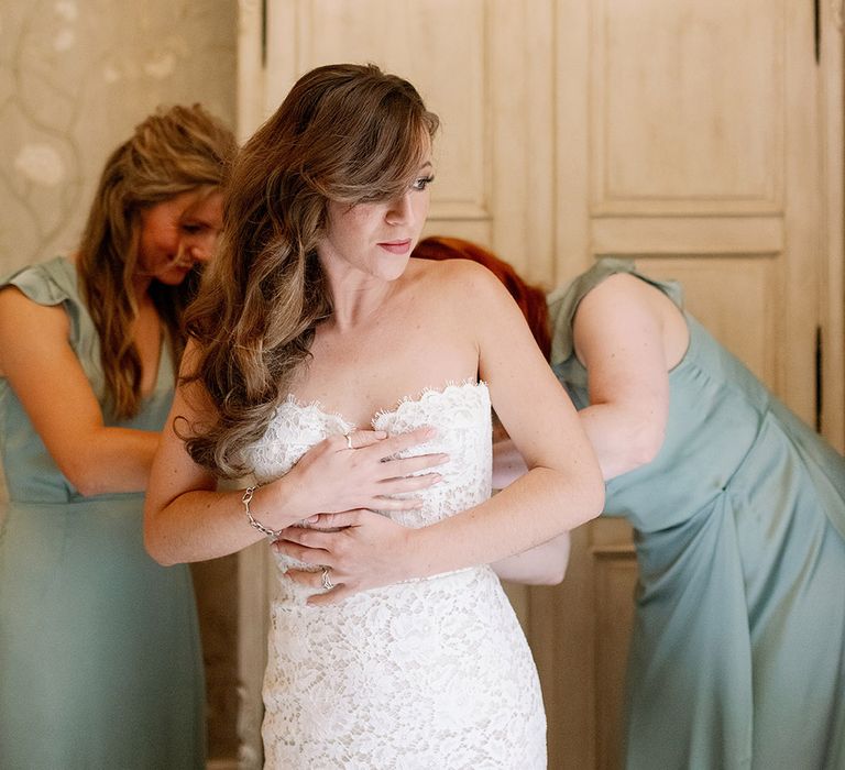 Bridesmaids in sage green dresses help the bride into her strapless lace wedding gown