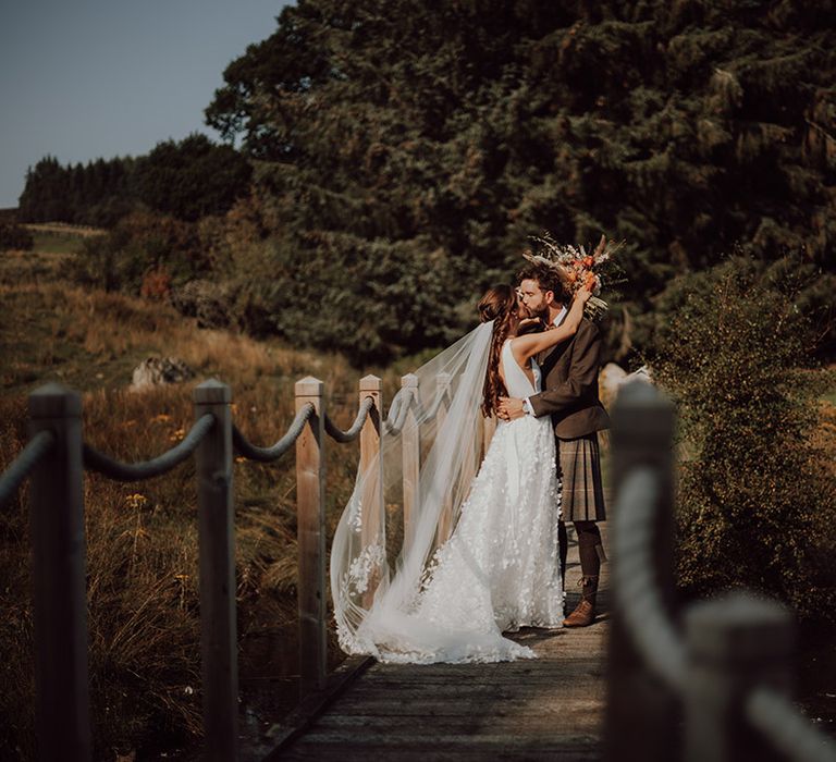 Bride in flower lace skirt wedding dress with matching veil kisses groom on bridge in traditional kilt 