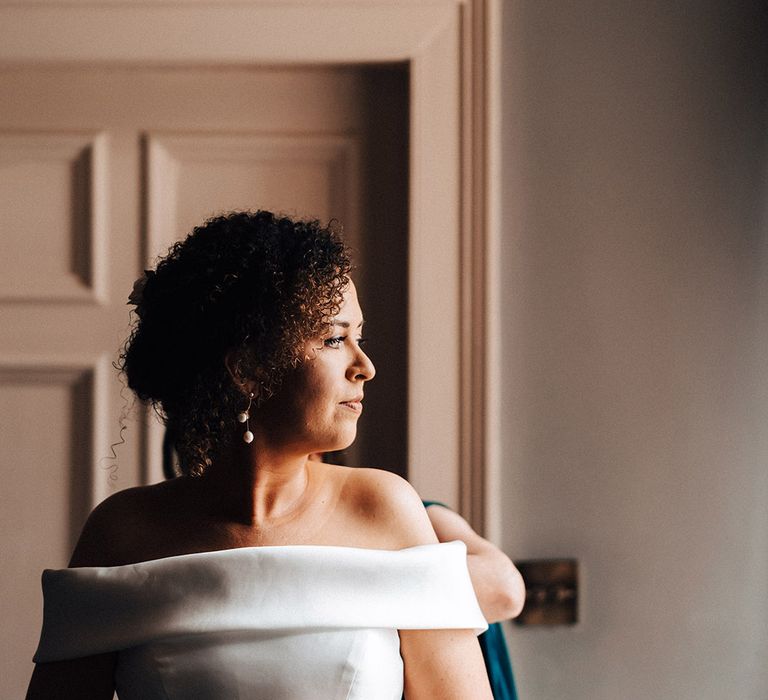 Bride is helped into pleated off the shoulder wedding dress with pearl earrings for wedding