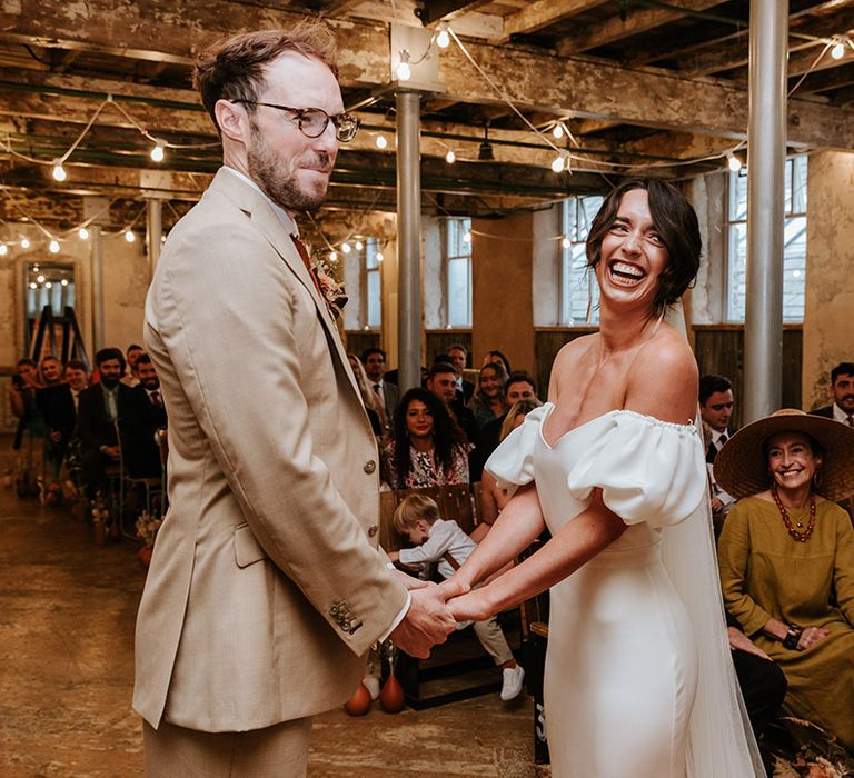 Groom in beige suit and brown glasses holds hands with the smiling bride in puff sleeve wedding dress at the altar