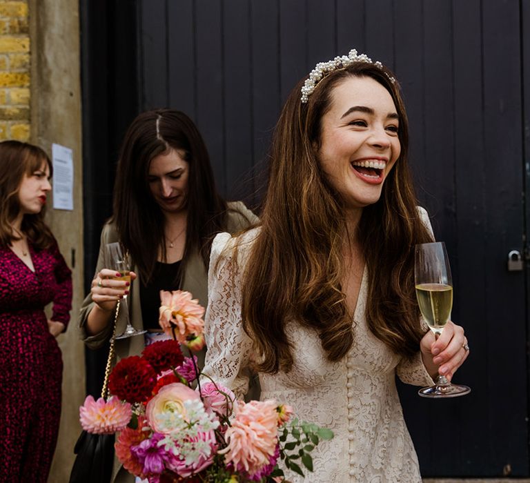 Smiling bride in lace Rico wedding dress with buttons and puffed sleeves holding drink and pink wedding bouquet 