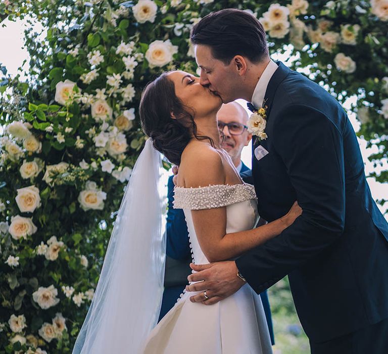 Groom in black tie has first kiss as a married couple with bride in sparkly wedding dress in front of flower arch at altar