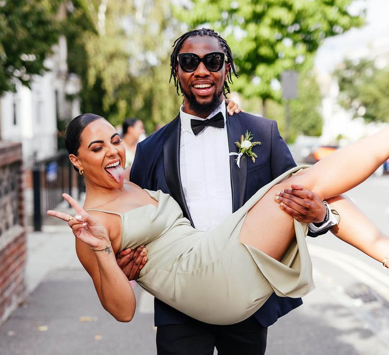 Groomsmen in blue and black tuxedo and large black sunglasses lifts bridesmaid in mint green dress in princess hold 