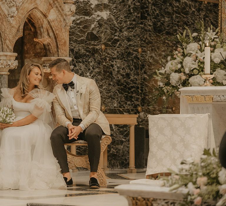 Bride and groom smile at each other as they sit at their wedding ceremony at Farm Street Church 