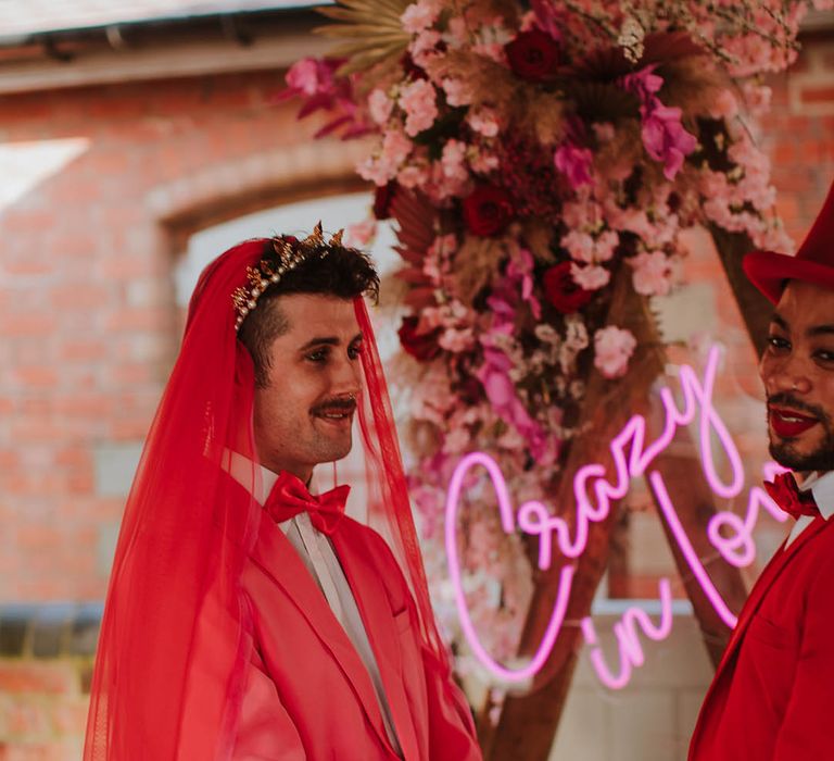 Stylish groom in a pink suit, gold crown and red veil holding hands with his groom at the altar 