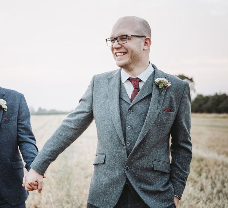 Grooms in three piece grey and dark blue suits hold hands together as they walk through fields wearing red ties and paper rose buttonholes