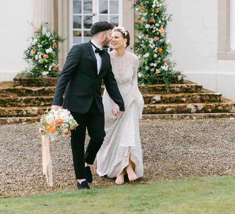 Bride in a beaded wedding dress with a chic up do and bridal headdress laughing with her groom in a tuxedo at Holesfoot Cumbria 