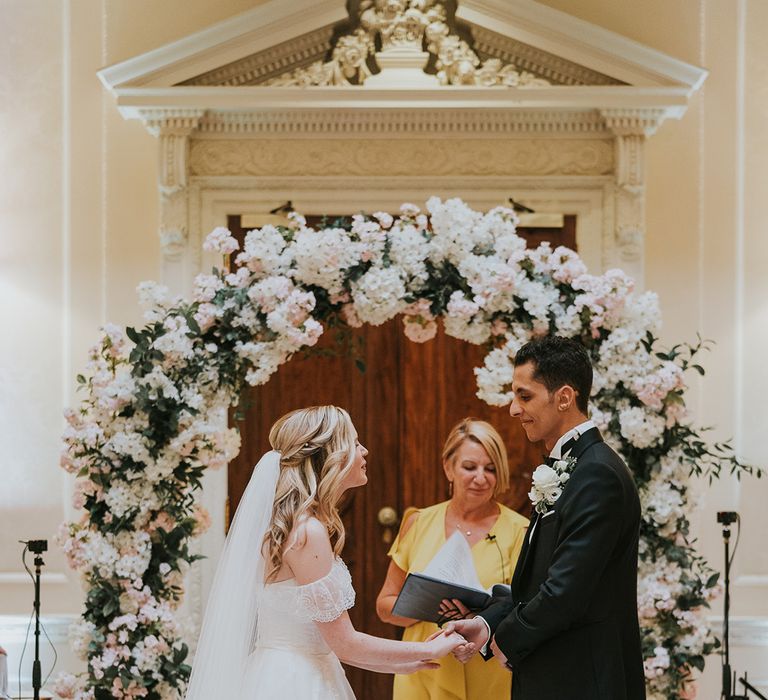 Bride and groom hold hands for their wedding ceremony in front of pink and white wedding flower arch 