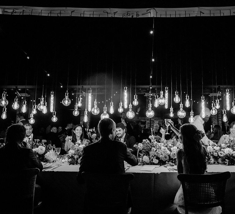 Black & white image of head table as Edison bulbs hang above bride & groom who cheers on their wedding day