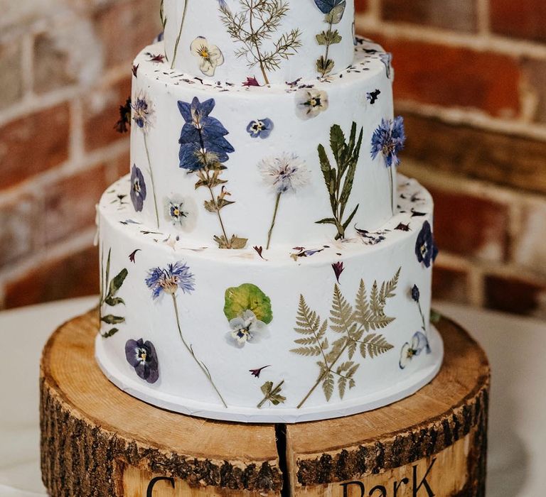 Three tier white frosted pressed flower wedding cake with wooden cake toppers of the bride and groom and their pets