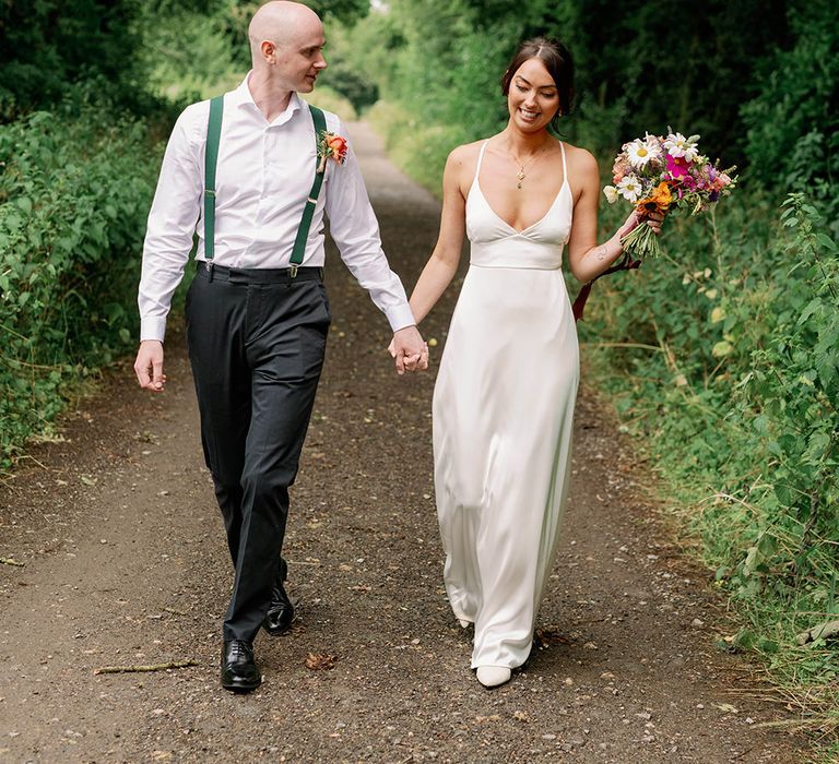 Bride walks with her groom whilst wearing DIY wedding dress in satin silk whilst holding brightly coloured floral bouquet 