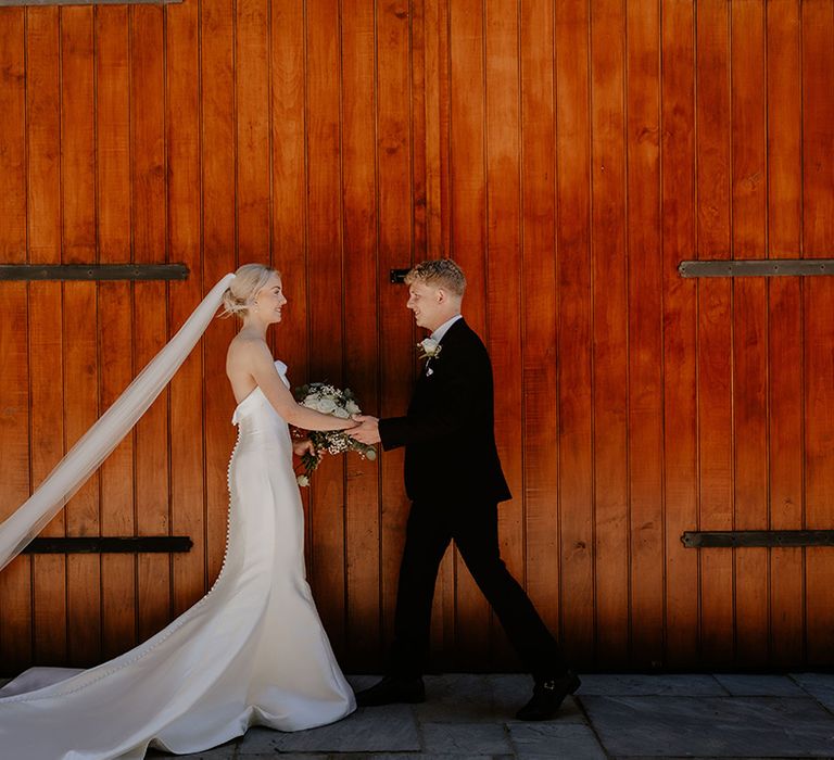 Bride in button back wedding dress with a train and veil with groom in black tie in front of wooden doors 