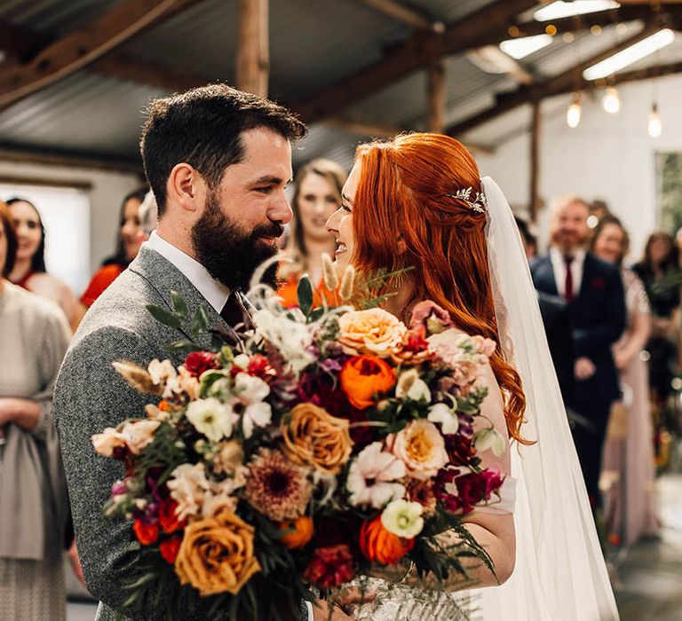 Bride and groom stand at the altar together as the bride shows off her large red and orange autumnal bouquet