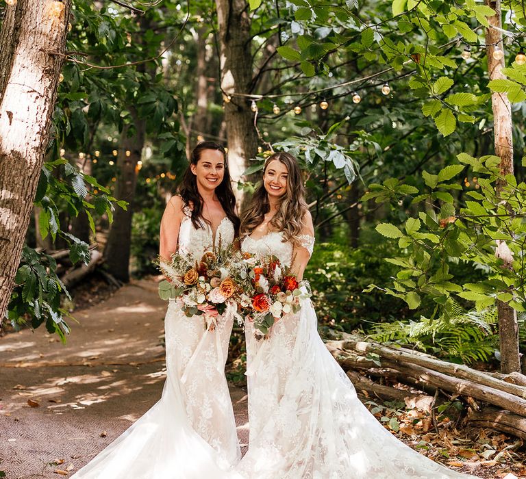 Brides stand together in boho lace gowns with warm toned autumn flower bouquets 