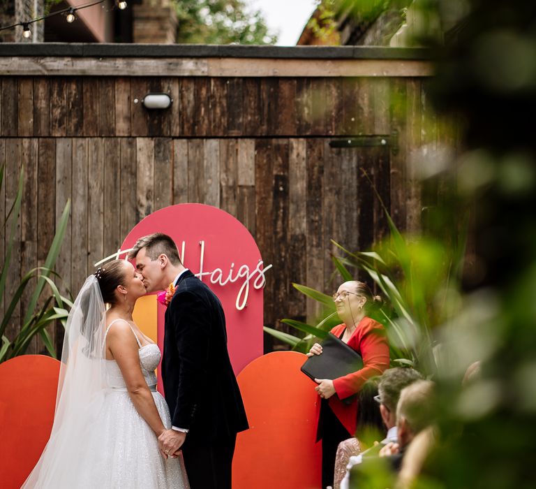 Bride & groom kiss in front of DIY altar with orange and pink theme with personalised neon sign 