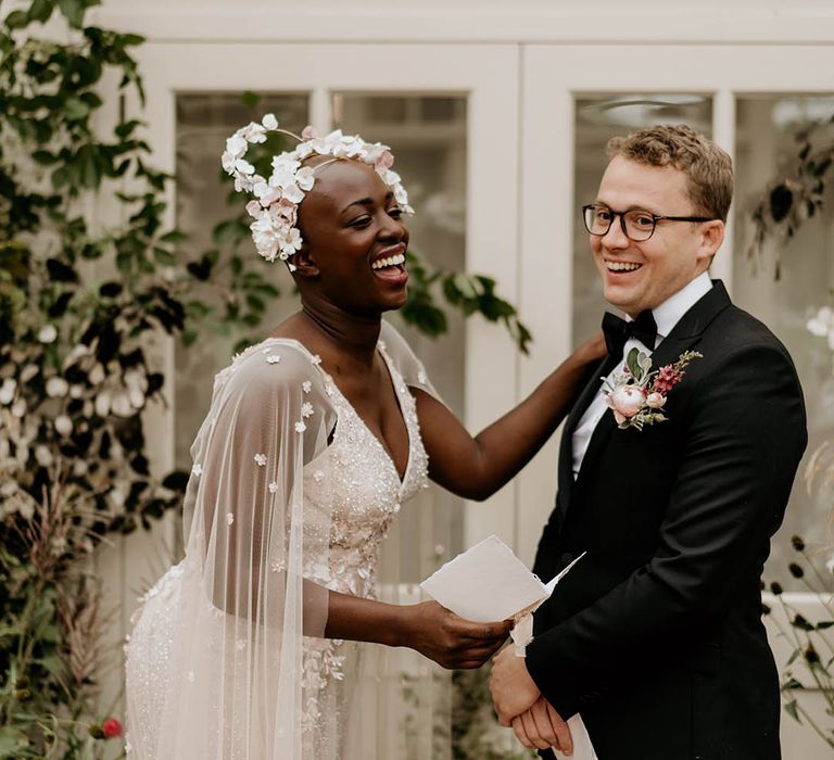 Bride wearing a flower headpiece and sheer cape laughs with groom in black tie for their ceremony 