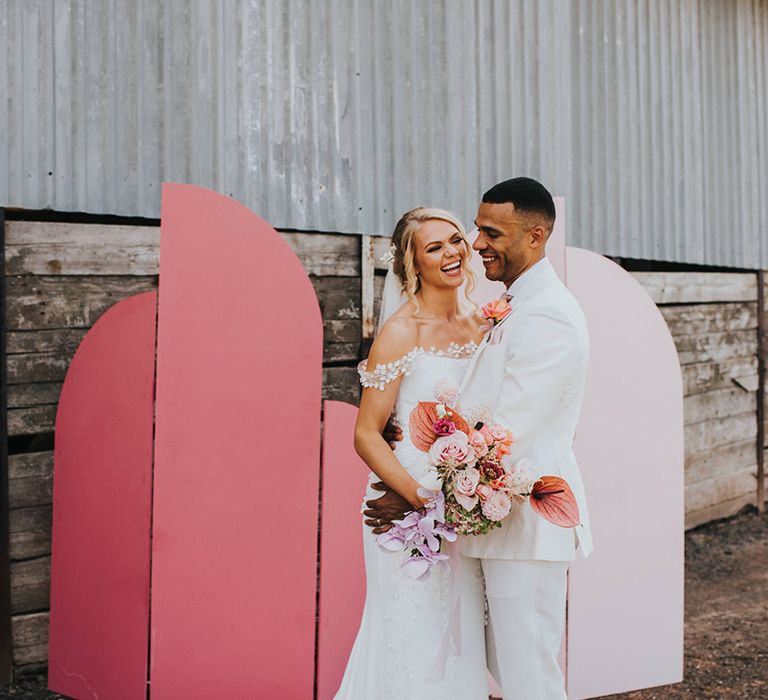 Groom embraces bride as they laugh together with a pink wedding screen background 