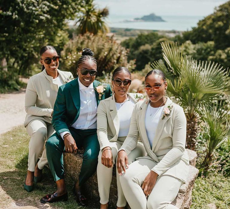 Black bride in a green suit sits with her bridesmaids who wear beige fitted suits with white T-shirts