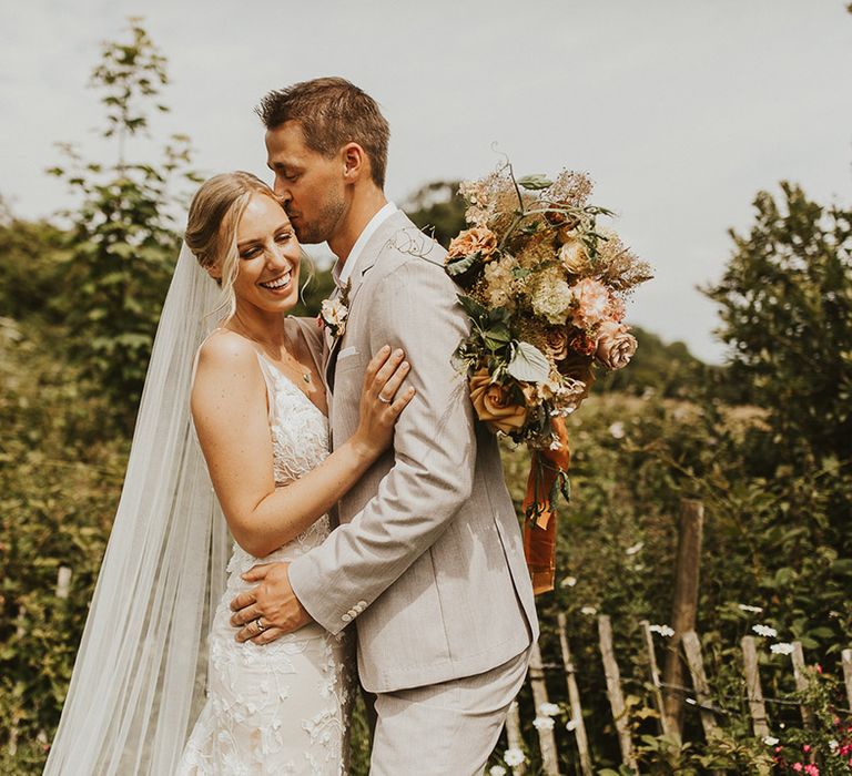 Bride holding a pink and rust coloured wedding bouquet embracing the groom as he kisses her on the head 