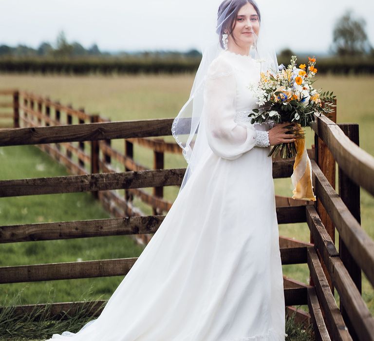 Bride in a vintage long sleeve high neck wedding dress holding a colourful blue, orange and yellow bouquet 